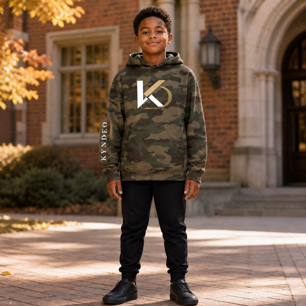 Forest Camo KYNDEO Youth Camo Hoodie on a Black boy model in a fall courtyard